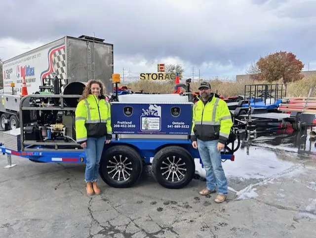 An image showing two workers in high-visibility safety vests standing next to a blue plumbing and drain service van.