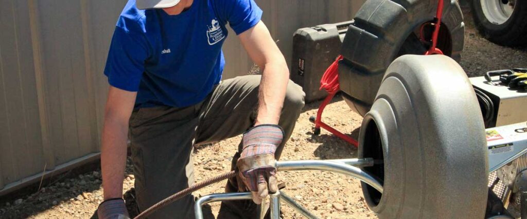 A person in a blue shirt working on a piece of equipment in a garage setting with various tools and equipment visible.