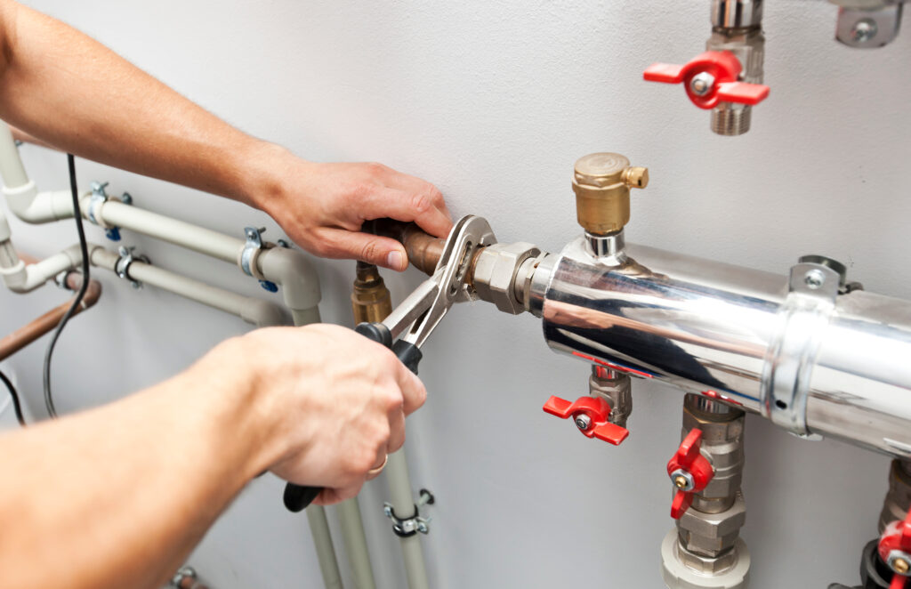 An image showing a person's hands working on a plumbing fixture, with various plumbing tools and pipes visible.