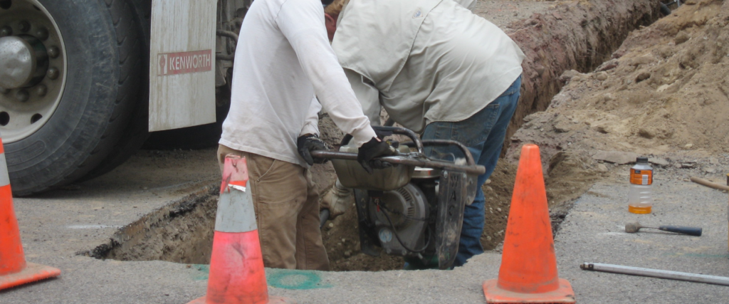 An image showing a construction worker using a tool to work on an underground pipe or cable, with traffic cones and construction equipment visible in the background.